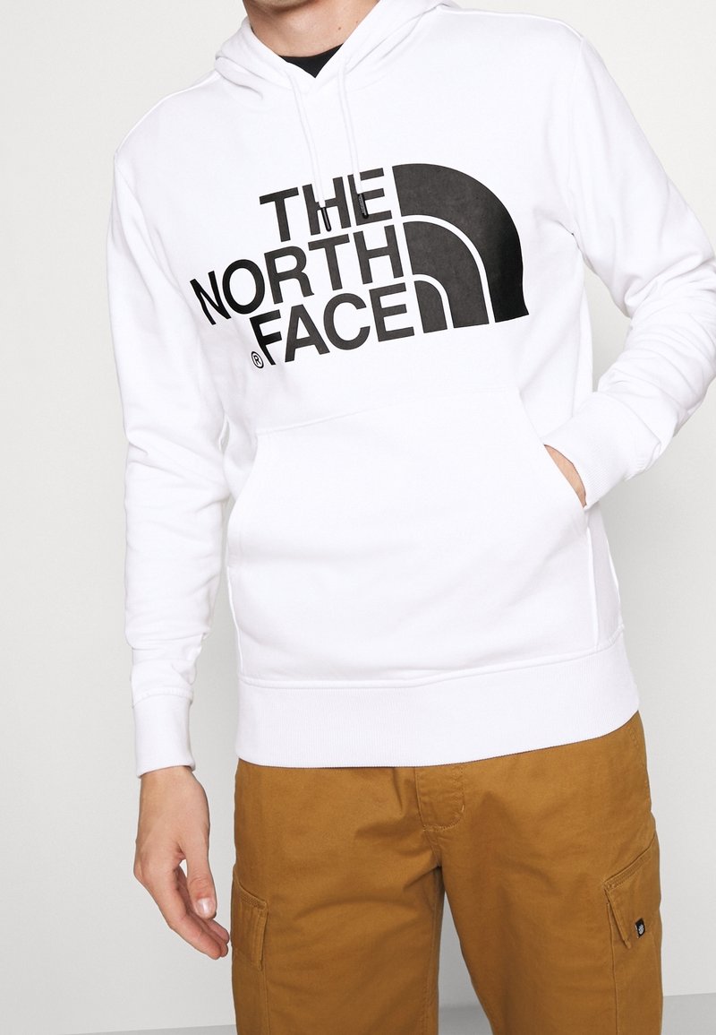 Persona che indossa una felpa bianca con un grande logo nero "The North Face" e pantaloni cargo marroni, con una mano in tasca della felpa.
