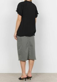 Blouse noire à manches courtes avec détails à volants, associée à une jupe kaki longueur genou avec ourlet brut, et des mules à talons noires.