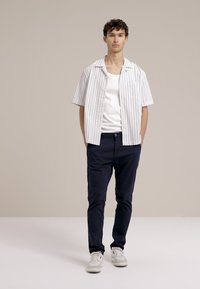 Cămașă albă cu mânecă scurtă și dungi albastre verticale, purtată peste un top alb ajustat. Pantaloni de marină și adidași albi completează ținuta.