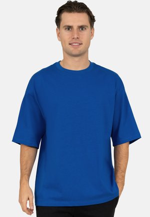 Uomo in piedi, indossa una maglietta blu semplice oversize a maniche corte e pantaloni neri, su sfondo bianco.