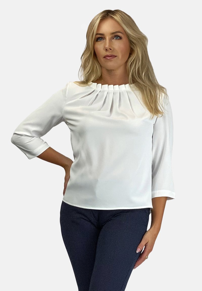 Biała bluzka z plisowanym dekoltem, rękawami o długości trzech czwartych i gładką fakturą. Noszona z ciemnymi spodniami w wzory.