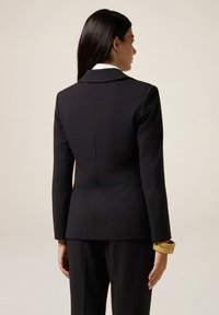 Blazer fitted nero, design strutturato, apertura posteriore singola, taglio slim, abbinato a pantaloni scuri, con un bracciale dorato al polso.
