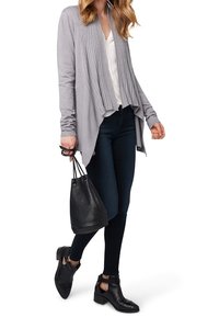 Ljusgrå ribbade cardigan med öppen front, ihop med en vit topp och mörka skinny jeans. Håller en svart bucket bag och har på sig ankelboots.