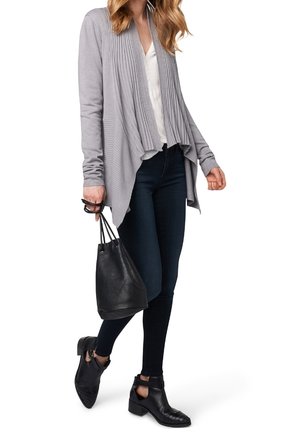 Cardigan a coste grigio chiaro con apertura frontale, abbinato a un top bianco e jeans skinny scuri. Porta una borsa a secchiello nera e indossa stivaletti.