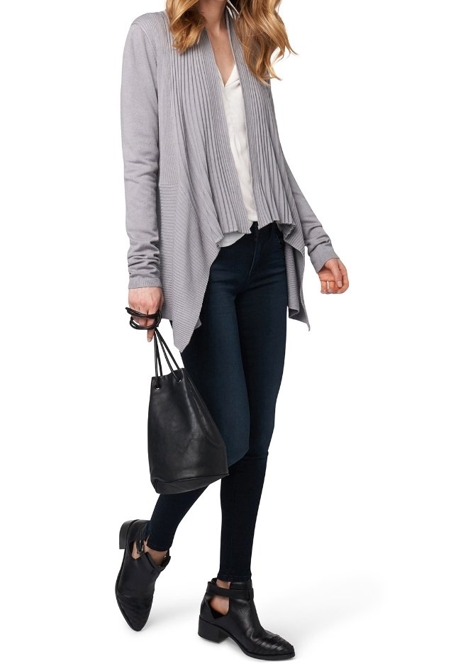 Ljusgrå ribbade cardigan med öppen front, ihop med en vit topp och mörka skinny jeans. Håller en svart bucket bag och har på sig ankelboots.