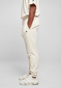 Pantalones jogger color crema, confeccionados en tejido suave, con bolsillos laterales, cintura con cordón y un pequeño logo negro en el muslo.
