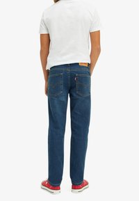 Blue denim jeans with a straight cut, featuring two back pockets, yellow stitching, and a leather logo patch on the waistband.