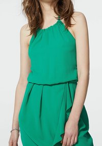 Zielona sukienka na ramiączkach z teksturowanej tkaniny, z marszczonym dekoltem i warstwowym, opadającym dołem. Prosty fason z luźną sylwetką.