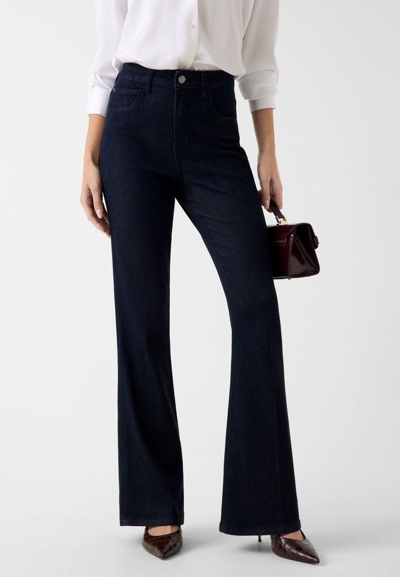 Jeans a zampa scuro con vita alta, tasche frontali e chiusura con bottone a contrasto. Il materiale ha una consistenza liscia di denim.