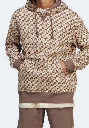 Homme portant un sweat à capuche beige et marron Adidas avec un motif géométrique du logo et un pantalon marron assorti, mains dans la poche du sweat.