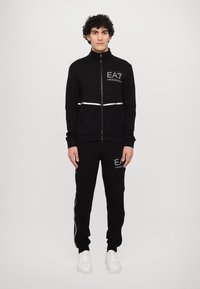 Black zip-up tracksuit jacket with a high collar and reflective stripe; paired with matching black pants featuring white side stripes and logos.