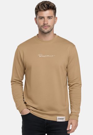 Mann trägt ein hellbraunes Langarmshirt mit weißem Schriftzug auf der Brust und einem Aufnäher nahe dem Saum, steht mit einer Hand in der Tasche vor schlichtem Hintergrund.
