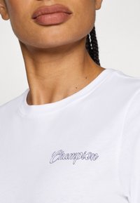 Witte katoenen t-shirt met een ronde halslijn en een klein paars "Champion" logo geborduurd aan de linkerkant. Gladde textuur.