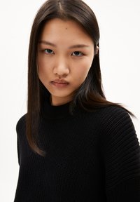 Schwarzer Strickpullover mit hohem Ausschnitt und strukturierter Stoffoberfläche. Eine Nahaufnahme betont das gerippte Muster und die Formung im Schulterbereich.