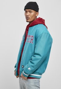 Teal varsity jakna z usnjenimi rokavi, vezenim napisom v maroon barvi, čez rdeč kapuco, bele črtaste manšete in črno beanie kapo.