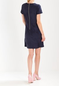 Robe bleu marine à manches courtes avec une coupe droite et un tissu texturé, dotée d'une fermeture éclair dorée visible dans le dos. Portée avec des talons hauts roses.