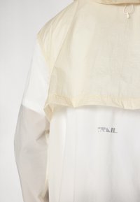 Giacca leggera di color crema con tessuto testurizzato, colletto a strati e la scritta "TRAIL" stampata in grigio sulla schiena.