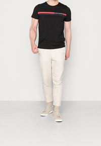 Camiseta negra de manga corta con una franja roja horizontal y un logo, combinada con pantalones tapered beige claro y zapatillas beiges.