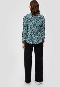 Langarm blouse in zwart, teal en wit met een bloemmotief. Heeft een ronde zoom en knoopdetails op de achterkant. Gecombineerd met zwarte wijde broeken.