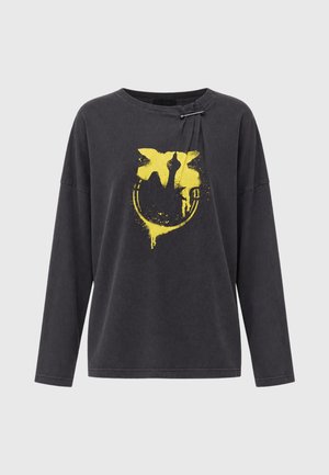 Femme debout portant un t-shirt noir à manches longues avec un motif jaune et un pantalon en cuir rouge, associé à des talons ouverts noirs sur fond blanc.
