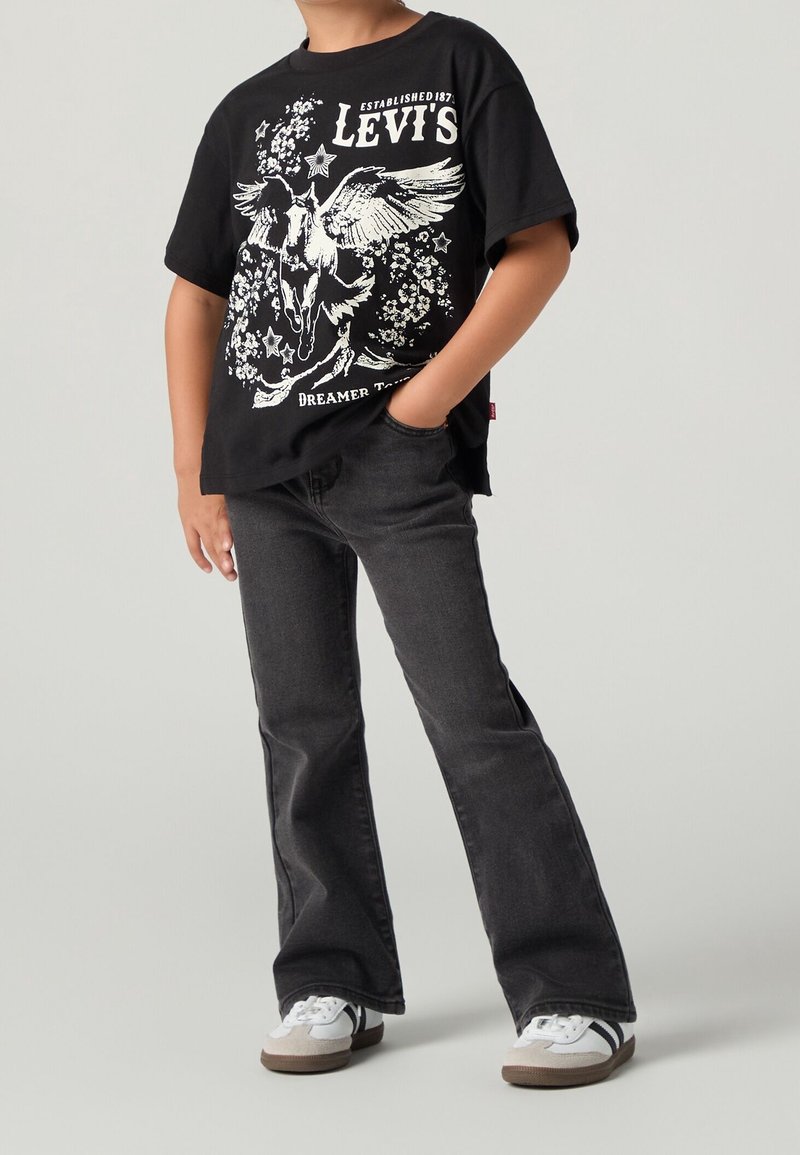 Enfant portant un t-shirt noir Levi's avec un motif blanc de Pégase, associé à un jean évasé foncé et des baskets blanches, debout avec une main dans la poche.
