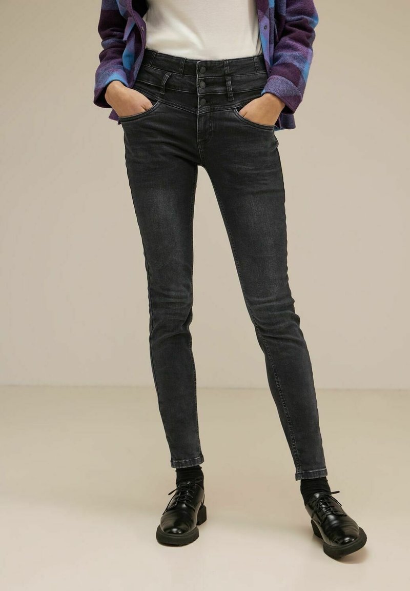 Personne portant un jean skinny noir taille haute avec trois boutons, des chaussures noires à lacets, un haut blanc et une veste à carreaux violette.