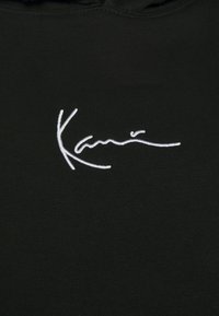 Čierna mikina s kapucňou, ktorá na oblasti hrudníka obsahuje biele vyšívané logo. Látka vyzerá mäkko, s hladkou textúrou.