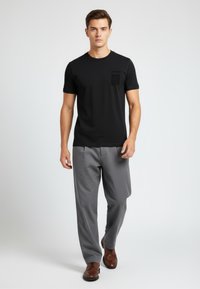 Schwarzes Kurzarm-T-Shirt mit einer strukturierten Tasche, kombiniert mit grauen Hosen und braunen Schuhen, vor einem neutralen Hintergrund.