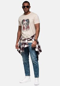 Beige T-shirt med et kraniegrafik og farverige accenter, blå jeans, hvide sneakers og en bundet ternet flannel skjorte.