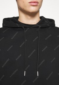 Sudadera negra con estampado de logo "SANDRO PARIS," con capucha ajustable y terminales plateados, hecha de una tela suave.