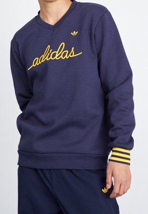 Mężczyzna ubrany w granatową bluzę Adidas z żółtym logo oraz dopasowane granatowe spodnie z żółtym emblematem Adidas.