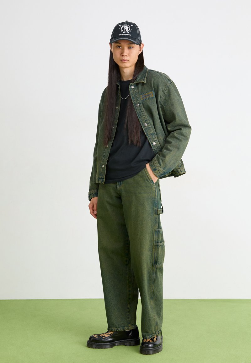 Giacca di jeans verde, t-shirt nera, pantaloni cargo verdi con tasche, stivali neri con un accento a stampa leopardata e un cappellino nero con un logo.