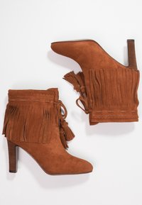 Stivaletti in suede marrone con tacco alto, caratterizzati da dettagli di frange, nappine e cuciture decorative lungo il bordo superiore.