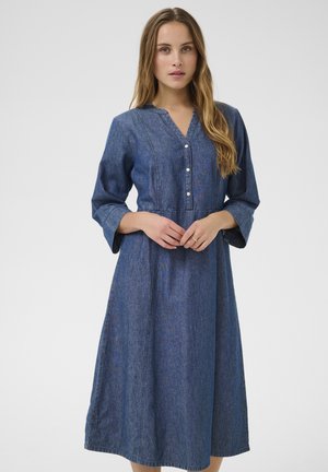 Denim ruha V-nyakkivágással, hosszú harangujjakkal és gombos elülső résszel. Simaság jellemzi, és a derékrésze ki van hangsúlyozva, amely egy bővülő szoknyába torkollik.