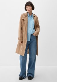 Vrouw staat met haar hand in haar zak, gekleed in een tan trenchcoat, lichtblauw shirt, wijdvallende blauwe jeans en zwarte schoenen tegen een witte achtergrond.