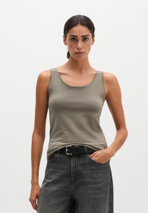 Vrouw met donker haar naar achteren gebonden, die een grijze mouwloze top draagt, gestopt in zwarte high-waisted jeans met een zwarte riem.