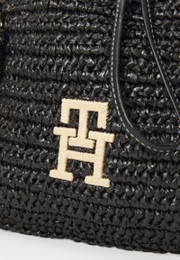 Sort vævet håndtaske med en struktureret overflade, prydet med et fremtrædende beige logo-design på fronten og sorte læderdetaljer.