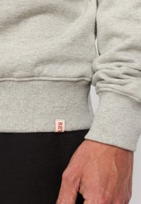 Grijze sweatshirt met geribbelde boorden en zoom, met een klein rood label. De stof heeft een zachte textuur, zichtbaar in een close-up detail.