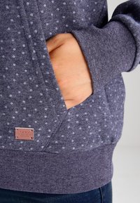 Tmavě šedá mikina s kapucí a světlými šedými puntíky, s přední kapsou a ribbovaným lemem. Na boku je viditelná malá kožená štítková značka.