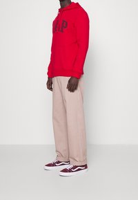 Sudadera roja con capucha y letras negras, pantalones anchos beige y zapatillas burdeos con detalles blancos, todo sobre un fondo blanco.