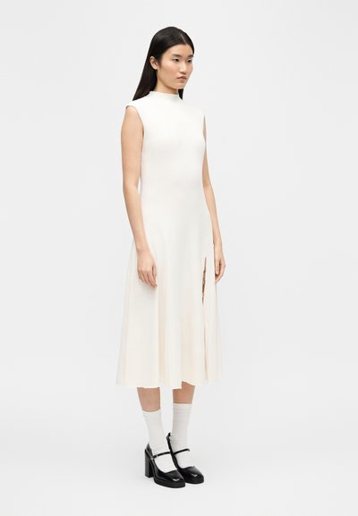 Femme aux longs cheveux bruns portant une robe midi blanche sans manches avec une fente à l'avant, des chaussettes blanches et des escarpins noirs style Mary Jane.