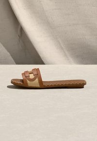 Καφέ επίπεδη σαγιονάρα με πλεκτό μπεζ λουράκι και διακοσμητικό ράψιμο κατά μήκος της σόλας, τοποθετημένη σε ανοιχτή επιφάνεια με φόντο από ύφασμα.