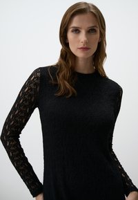 Schwarzes Kleid aus strukturiertem Stoff mit langen Spitzenärmeln, taillierter Passform und runder Ausschnitt. Der glatte Oberteil geht in kunstvolle Muster über.