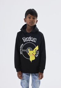 Črna kapuca s sprednjim grafičnim motivom Pikachuja in Pokéballa, okrašena z belim besedilom: "Gotta Catch 'Em All." Mešanica bombaža.