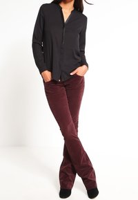 Blusa nera a maniche lunghe con collo a V e dettaglio dei bottoni, abbinata a pantaloni a zampa d'elefante in corduroy bordeaux scuro e calzature nere.