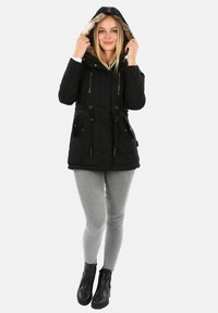 Frau trägt eine schwarze Kapuzenparka mit Fellbesatz, hellgraue enge Jeans und schwarze Knöchelstiefel, steht vor weißem Hintergrund.
