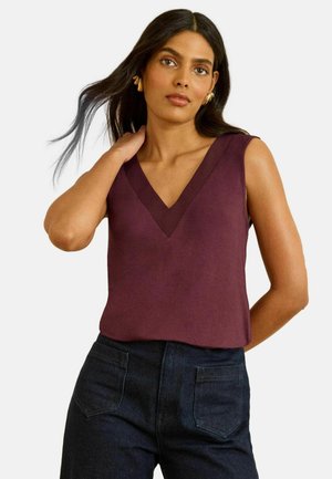 Ärmelloses V-Ausschnitt-Oberteil in tiefem Burgunderrot, aus glattem Stoff gefertigt. Kombiniert mit dunklen Denimhosen mit Gesäßtaschen. Einfaches Design.