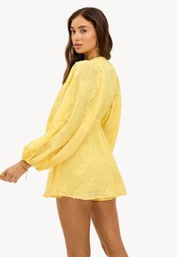 Robe jaune brodée avec des manches longues bouffantes, une coupe ample et des motifs floraux complexes sur un tissu texturé, atteignant la mi-cuisse.