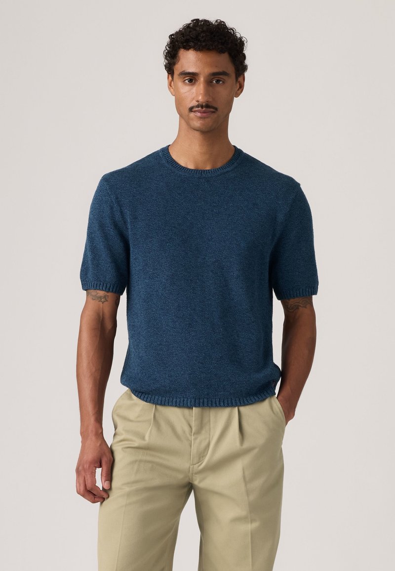 Mann mit kurzen lockigen Haaren und Schnurrbart, trägt einen blauen Kurzarmpullover und beige Hosen, steht mit einer Hand in der Tasche.
