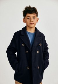 Poiss lühikeste pruunide juustega kannab tumesinist kahekordse nööpidega mantlit sinise särke üle, seistes käed taskutes, lihtsa tausta ees.